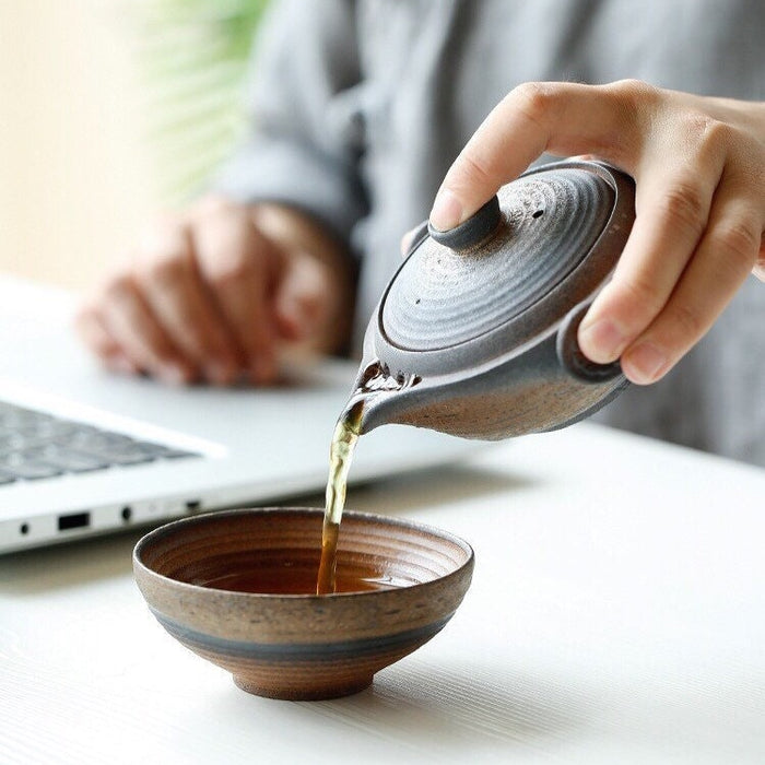 Person pouring tea from a ceramic teapot into a bowl on a desk with a laptop in the background.
