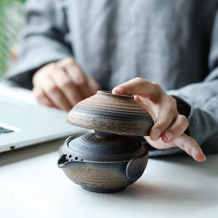 Person holding a ceramic teapot on a desk with a laptop in the background