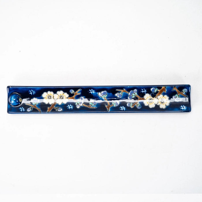 A long, narrow rectangular ceramic incense holder with a glossy deep blue background, decorated with white cherry blossom flowers and brown branches, on a plain white background.
