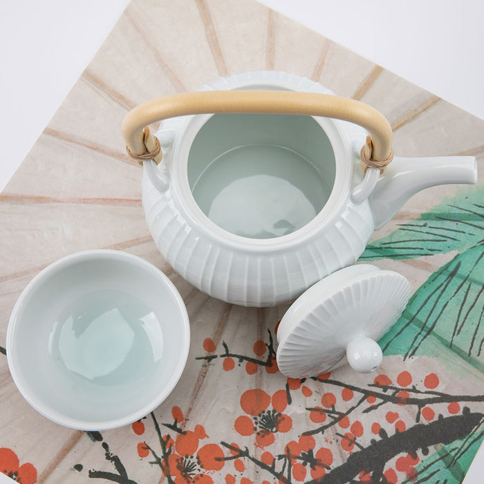 White ceramic teapot with a wooden handle and a matching cup on a decorative mat with floral patterns.