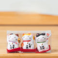 Three small ceramic cat figurines in packaging on a wooden surface