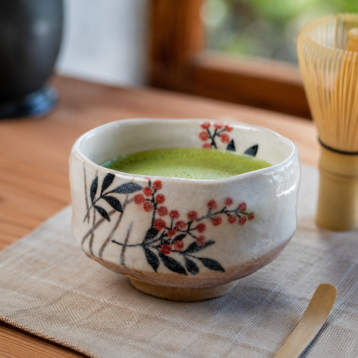 A handmade ceramic matcha bowl filled with green matcha tea, decorated with black leaves and red berries on a cream-colored glaze, resting on a woven placemat with a bamboo whisk in the background.