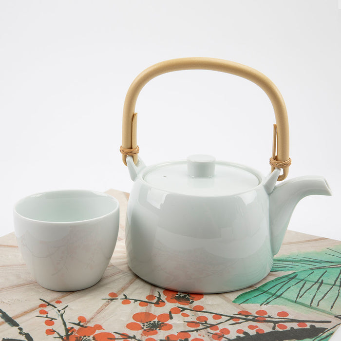 White ceramic teapot with a wooden handle and a matching cup on a decorative tablecloth.