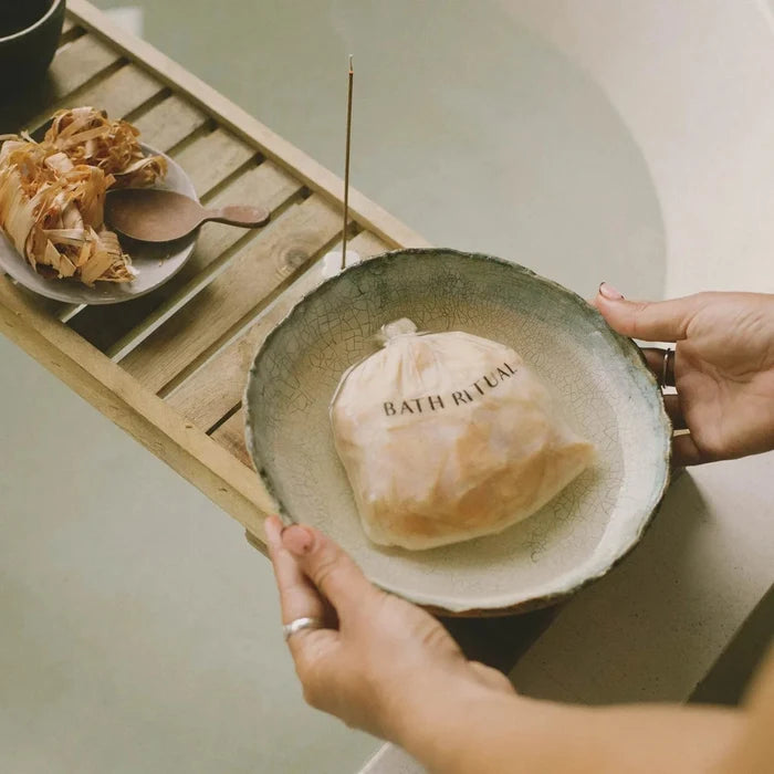Person holding a ceramic bowl with a bag labeled 'Bath Ritual' on a wooden tray.