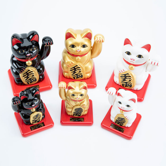A collection of solar-powered Maneki Neko figurines in various colors including black, gold, and white, displayed on a red base, waving their paws.